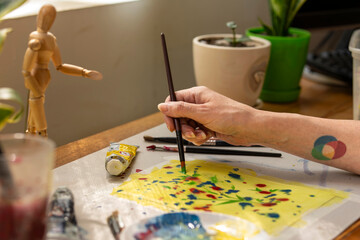 Female artist hand holding a brush while creating colorful abstract dots and strokes on yellow painted paper.
