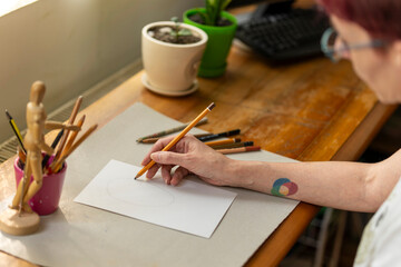 Over the shoulder view of a woman sketching with a pencil on white paper at a wooden desk with art...