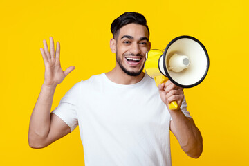 Excited arab guy speaking loud and raising hand up, using megaphone on yellow studio background....