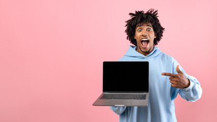 Shocked black teen guy pointing at laptop with blank screen on pink studio background, mockup for website design. Funky Afro adolescent demonstrating PC with space for your online advertisement