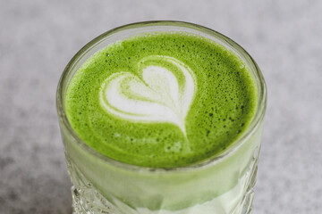 A glass of creamy matcha latte with heart-shaped latte art on top. The vibrant green drink is...