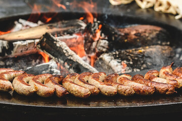 Grilled sausages cooking on an open fire pit with wood logs and glowing embers. The rustic setup suggests outdoor cooking or a traditional barbecue