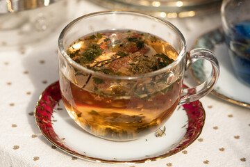 Glass cup filled with brown hot herbal tea, infused with dried flowers and herbs, placed on a decorative saucer. A warm, cozy beverage often enjoyed for its soothing and healthful properties