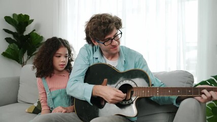 Caucasian father teaching cute daughter about playing guitar while sitting on sofa. Cute student girl looking at his dad playing guitar while learning about music. Family recreation concept. Pedagogy. - Powered by Adobe