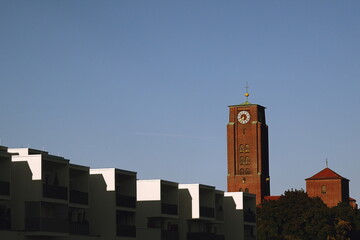 Red brick church tower with a clock rising above modern white apartment buildings under a clear blue sky.