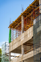 Upward perspective of new urban building development showcasing raw concrete elements, elaborate metal scaffolding, wooden formwork for floors, and protective netting against vivid blue sky.