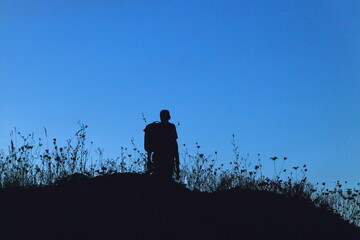 Silhouette of a lone hiker with a backpack standing on a hill at twilight, surrounded by wild grasses against a deep blue evening sky.