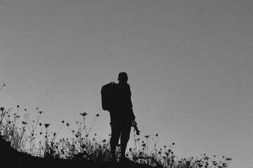 Silhouette of a lone hiker with a backpack standing on a hill at twilight, surrounded by wild grasses against a deep blue evening sky.