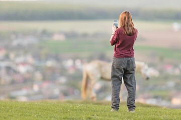 A woman is seen from the back, using her mobile phone to take a picture of a horse in the meadow during dusk. A blurred village is in the background