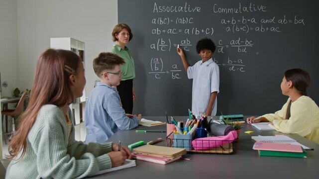 Young African American student at blackboard explaining associative and commutative properties to engaged classmates in classroom