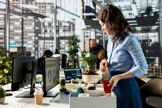 Optimistic excited person unpacking her office belongings at the new job, settling in at workstation and feeling enthusiastic about joining the corporate team. Female newcomer unboxing things.