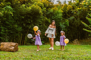 Fototapeta premium Mother smiles at twin daughters holding balloons during outdoor birthday, as one girl looks upset.
