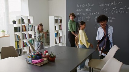 Teacher standing near chalkboard of equations while middle-school students unpacking their belongings in classroom - Powered by Adobe