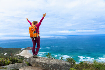 Athletic woman on mountain trail, standing on rock with backpack and ocean view. Outdoor training, trail running, hiking for strength, health and fitness motivation. Powerful coastal adventure moment.