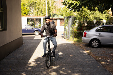 Naklejka premium Black delivery worker riding a bike in the driveway carrying large thermal bag, navigating urban streets on the delivery bike to bring meal order to client. Express shipping service.