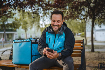 Obraz premium Delivery man using smart phone app and smiling sitting on bench with thermal bag