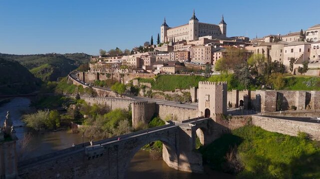 Drone view flying along the Tagus River towards the ancient Puente de Alc&aacute;ntara stone bridge and the towering Alc&aacute;zar de Toledo fortress, dominating the historic Spanish skyline.