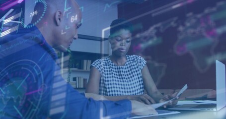 Reviewing documents and typing on laptop two colleagues at office desk, with holographic charts