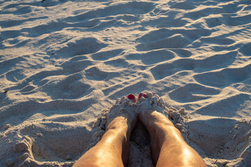 Two sets of toes poking out of fine white sand. The female's feet have manicured pink toenails on tan legs. There are footprints in the sand surrounding the woman's feet. The sun is glowing yellow.