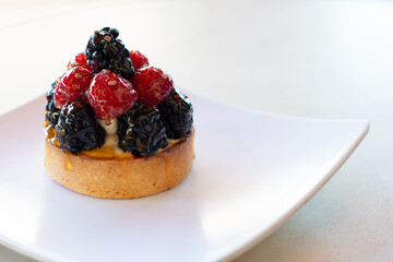 Fruit-filled French custard tart. The topping has fresh red raspberries and blackberries on a round almond baked crust. The sweet dessert is on a white square plate on a table for sale at a bakery. 