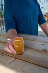 A small, clear sample beer glass filled with sour ale on a picnic table at a microbrewery. The fruit beer has froth on the top around the rim. A man is wearing a blue shirt, holding a beer glass