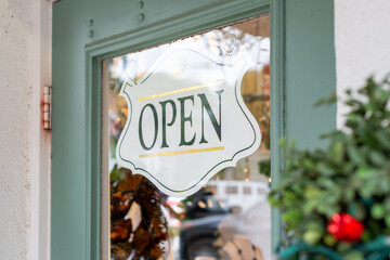 A white wooden open sign with green text and gold stripes hangs in the window of a shop door.The green wooden door is closed.The business is decorative in the background. A rose bush is near the sign.