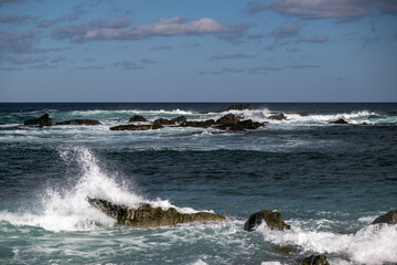 A rugged coastline of rock, cliffs, boulders along the shoreline in Newfoundland. The sky is deep blue with white clouds along the horizon of the ocean. The water has waves rolling on the rocky shore.