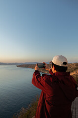 Obraz premium Young man taking a photo with his cell phone of the seascape at an incredible sunset, concept of tourism, technology, inspiration, and lifestyle