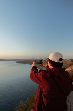 Young man taking a photo with his cell phone of the seascape at an incredible sunset, concept of tourism, technology, inspiration, and lifestyle