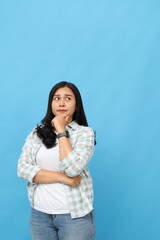 Fototapeta premium Thoughtful young Asian woman with hand on chin looking up thinking or contemplating something, standing isolated on blue background.