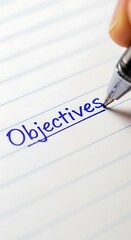 A close up of a pen writing the word objectives in blue ink on lined white paper at a slight angle