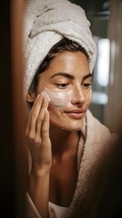 Woman applying facial mask in bathroom mirror wearing towel on head and bathrobe close-up beauty skincare routine self-care wellness