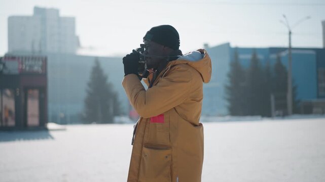 round view of shutterbug in yellow parka and beanie capturing snowy urban landscape with vintage camera, strap on neck, cars and buildings blurred in background under soft cold winter daylight
