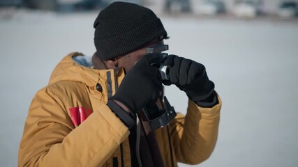 side view of black man peering through camera viewfinder and adjusting focus dial while reviewing shot on LCD display against snowy urban scene with blurred trees and parked cars under winter sun