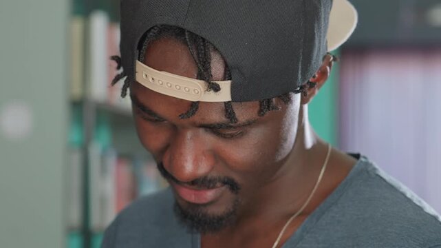Close up of young man wearing backward cap smiling softly while tilting head down, showing relaxed expression and subtle dimples, in warm indoor setting with soft background