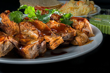 A close-up image of BBQ pork ribs with cilantro on top, with corn tortillas, Mexican rice, and guacamole in the background.