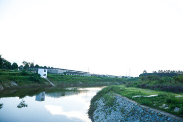Fototapeta premium Industrial buildings along grassy riverbank with water reflections and natural landscape