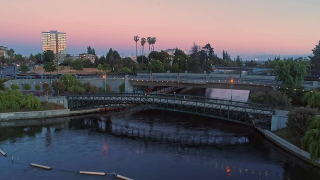The Los Angeles River flows under bridges at dusk in Glendale, California, USA. The river is a concrete channel with water flowing through it. People walk across the bridges.