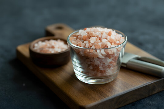 Himalayan Pink Salt Presentation: A close-up shot of Himalayan pink salt crystals in a glass and wooden bowl. The crystals sit atop a wooden cutting board.  A metal spoon is beside the glass. - Powered by Adobe