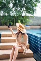 Young Hispanic Gen Z Woman Using Smartphone to Update Social Media by Resort Pool During Summer Vacation.