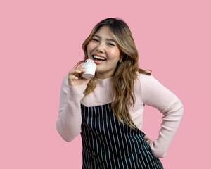 Young asian woman with an apron in celebratory or festive mood holding cupcake, decorated with white frosting and chocolate chip. Portrait on pink background with studio light, Isolated.