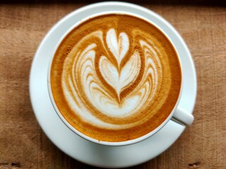 A beautifully crafted latte with intricate foam art, served in a white cup on a wooden surface, showcasing rich coffee tones and creamy textures.