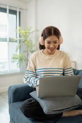 A young woman wearing headphones sits comfortably on the sofa, smiling while working on her laptop. She looks focused and happy, representing modern lifestyle and remote work.