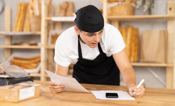 Young bakery owner in black uniform leaning on wooden counter surrounded by baked goods, using phone while examining paperwork with puzzled, worried expression, uncovering problems with order details - Powered by Adobe