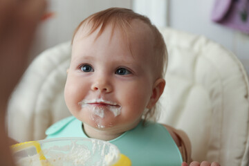 Little baby in bib eats with a spoon on his own while sitting at a feeding table