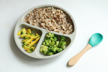 Silicone plate divider for feeding small children with buckwheat porridge, pieces of avocado and broccoli. Healthy food for baby
