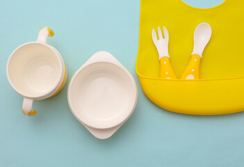 Silicone baby bib with bowls and spoons on blue pastel background. Accessories for feeding a small children. Top view