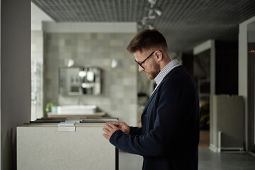 Fototapeta premium Caucasian young adult man with beard and glasses examining ceramic tile samples in showroom, standing in profile and focusing on surface texture, modern interior visible in background