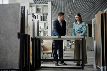 Caucasian man and Caucasian young adult woman examining large tile samples together in modern showroom, both standing and discussing material options for interior design project