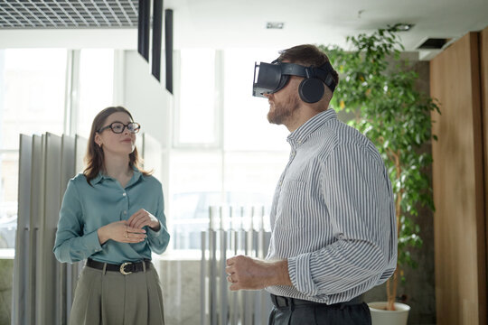 Caucasian man wearing virtual reality headset standing indoors while young woman with glasses observing him, both engaged in technology demonstration